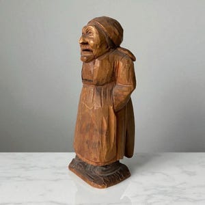 Peut inclure: Figurine en bois sculpté à la main représentant une personne âgée portant un couvre-chef et un long manteau. La figurine est posée sur une base rectangulaire. Le bois est de couleur marron chaud avec des détails sculptés.