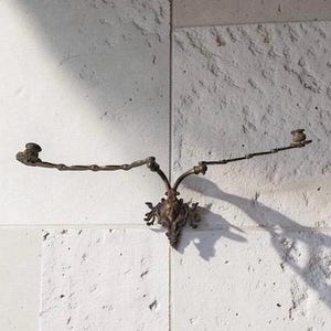 Könnte beinhalten: Ein antiker Wandleuchter aus Bronze mit zwei Kerzenhaltern. Der Leuchter hat ein verziertes, dekoratives Mittelstück und zwei nach außen gerichtete Arme. Der Leuchter ist an einer strukturierten, hellfarbigen Wand montiert und wirft einen Schatten.