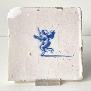 May include: A square ceramic tile with a white glazed surface featuring a network of fine cracks. Centered on the tile is a blue painted graphic of a winged figure holding a long object. The edges of the tile show signs of wear and chipping.