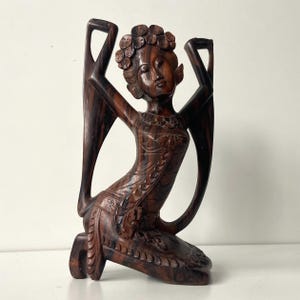 Può includere: Una scultura in legno inginocchiata di una donna con le braccia alzate. L'intaglio in legno marrone scuro presenta dettagli intricati, tra cui accenti floreali nei capelli e negli abiti. La figura ha un'espressione serena.