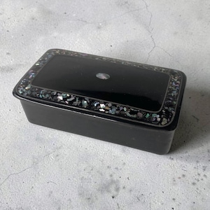 May include: A rectangular black box with a smooth, glossy finish. The lid features a central oval inlay and a wide border filled with small, iridescent, multi-coloured crushed shell fragments that shimmer with blue, green, and purple tones.