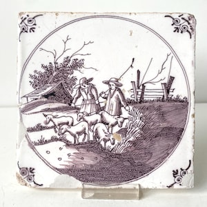 May include: A square, antique tile with a pastoral scene in shades of purple and white. The image depicts two figures herding sheep near a fence and a small building. Decorative floral corner accents complete the design.