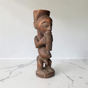 Può includere: Una statuetta in legno marrone intagliata a mano che raffigura una figura in piedi con una barba prominente, occhi a mandorla e testa piatta. La scultura presenta una superficie strutturata con venature del legno visibili su base circolare.