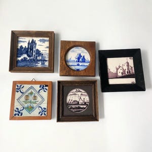 May include: Five framed ceramic tiles with various designs. The tiles feature images of buildings, landscapes, and floral patterns in blue, black, and white. The frames are made of wood in various shades of brown.
