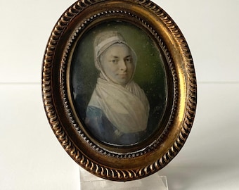 May include: Oval portrait miniature of a woman wearing a white cap and a white scarf, framed in a gold oval frame.