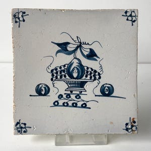 May include: Square ceramic tile with a white background and a blue fruit basket design. The design includes a pear, grapes, and two round fruits. Blue floral accents are in each corner.