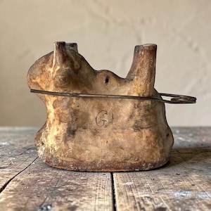 Puede incluir: Un objeto de madera antiguo, de color marrón claro y desgastado, con dos protuberancias verticales y un asa de alambre. El objeto tiene una marca circular con la letra "G" grabada en la parte delantera. La madera muestra signos de edad y desgaste.