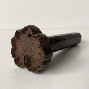 以下が含まれることがあります： 表面がざらざらとしていて小さな穴が開いた、花形のヘッドを持つダークブラウンの風化した金属製スタンプ。スタンプには、花形の金属ヘッドの背面から伸びる長い円筒形のハンドルが付いています。