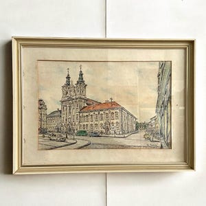 Puede incluir: Pintura a la acuarela enmarcada de una iglesia europea con dos campanarios y un tejado de tejas rojas. La obra de arte representa una escena callejera con coches antiguos y edificios. El marco es de color crema claro.