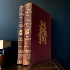 Puede incluir: Tres libros antiguos con lomos de cuero. Dos libros son marrones con letras doradas y patrones decorativos. El tercero es burdeos con un emblema dorado. Los libros están sobre una superficie de madera oscura.