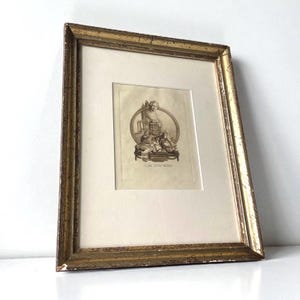 Op de afbeelding: Een antieke ingelijste prent met een goudkleurige, verweerde houten lijst. De prent toont een gedetailleerde zwart-wit illustratie van klassieke figuren in een ovale omlijsting. De prent is geplaatst op een crèmekleurige passe-partout.