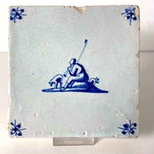 May include: Square ceramic tile with a light blue background and a blue illustration. The illustration depicts a person playing a wind instrument, with an animal nearby. Floral designs are in each corner.