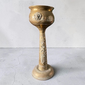 Pode incluir: Um vaso de pedestal em latão polido com uma taça larga e arredondada, apresentando um relevo de cabeça de leão com um anel. O caule cilíndrico fino e a base circular estão decorados com padrões intrincados de flores e volutas em relevo.
