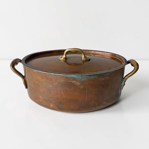 May include: A copper cooking pot with a lid and two side handles. The pot has a round shape and a tarnished patina. The lid has a brass handle, and the side handles are also brass. The pot is approximately 12 inches in diameter.