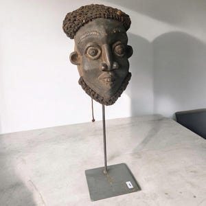 May include: A dark brown wooden mask depicting a human face with a braided hairstyle. The mask is mounted on a black metal stand with a square base.