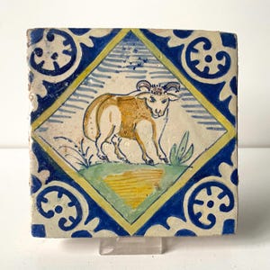 May include: Square ceramic tile with a hand-painted design. The central image features a tan and white ram within a yellow and blue diamond. The tile has a blue and white floral border. The tile is likely antique.