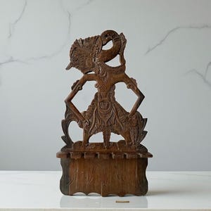 Op de afbeelding: Een gedetailleerde, bruine houten sculptuur van een traditionele Indonesische Wayang Kulit-pop. De pop is ingewikkeld gesneden met sierlijke details, staand op een rechthoekige basis. De figuur heeft een kenmerkende hoofdtooi en uitgestrekte armen.
