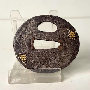 Op de afbeelding: Een ronde, donkerbruine metalen tsuba, een Japanse zwaardbeschermer, met twee openingen en gouden bloemaccenten. De tsuba wordt gepresenteerd op een transparante standaard, die het ingewikkelde ontwerp en de verouderde uitstraling benadrukt.