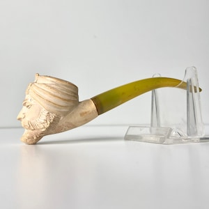 May include: A white ceramic pipe with a sculpted head wearing a turban. The pipe has a yellow stem.