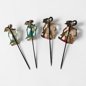 Op de afbeelding: Vier traditionele Indonesische schaduwpoppen, elk met ingewikkelde ontwerpen en bevestigd aan zwarte staven. De poppen hebben een verscheidenheid aan kleuren, waaronder groen, bruin, rood en goud, en staan tegen een witte achtergrond.