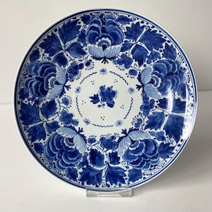 May include: A round ceramic plate featuring a white base with intricate blue floral patterns. The design includes large, detailed blue flowers and leaves arranged around the rim, with a smaller cluster of berries centered in the middle.
