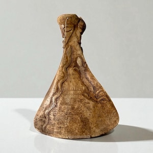 Op de afbeelding: Een bruin gekleurde, gebeeldhouwde sculptuur met een uitlopende basis en een smalle hals. De sculptuur heeft ingewikkelde geëtste ontwerpen, waaronder slangachtige figuren en geometrische patronen. De bovenkant van de sculptuur heeft een gestileerd menselijk gezicht.