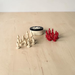 Pode incluir: Uma coleção de peças de xadrez e um pequeno recipiente redondo. As peças de xadrez são em duas cores: branco sujo e vermelho. O recipiente é branco sujo com uma tampa preta.