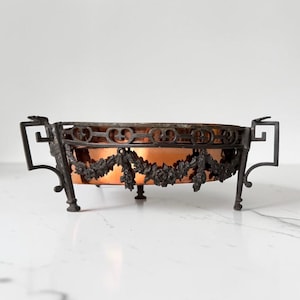 Puede incluir: Un cuenco redondo de cobre dentro de un marco de metal oscuro con guirnaldas florales ornamentadas y asas geométricas. El cuenco se apoya sobre pequeños pies y muestra un interior metálico pulido que contrasta con el exterior de metal oscuro.