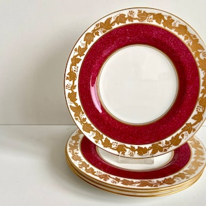 Puede incluir: Tres platos apilados con un diseño rojo y dorado. Los platos tienen un centro blanco y un borde dorado con un patrón de vid.