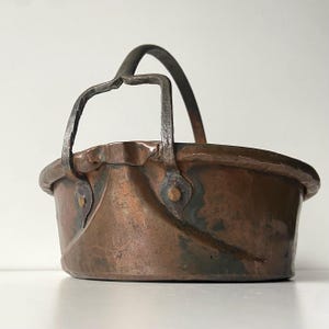 May include: A small, antique copper bucket with a dark, curved metal handle. The bucket has a rounded rim and is riveted with metal fasteners. The copper has a rich, aged patina with areas of green oxidation, suggesting its age and use.