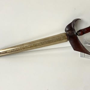 Könnte beinhalten: Ein goldfarbenes Keris, ein traditionelles indonesisches Dolch, mit einem Holzgriff und einer gebogenen Klinge. Die Klinge ist mit komplizierten Mustern verziert.