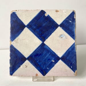 May include: Square ceramic tile with a repeating pattern of blue and white diamonds. The tile has a slightly aged appearance, with some wear and tear visible on the edges. The blue color is a deep, saturated hue.