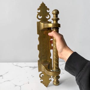 Può includere: Maniglia per porta in ottone dorato ornata, con placca posteriore decorativa e maniglia attorcigliata. La maniglia è tenuta da una persona. L'ottone ha una calda tonalità dorata e presenta dettagli intricati.