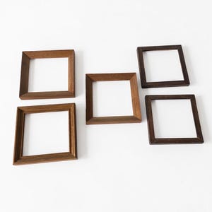 Può includere: Cinque cornici quadrate in legno di diverse tonalità di marrone disposte su sfondo bianco. Le cornici hanno un design semplice e minimalista, con venature del legno visibili. I centri vuoti suggeriscono che siano per esporre foto o opere d'arte.
