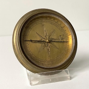May include: A round brass compass featuring a gold-toned face with an engraved star-shaped directional marker and a thin horizontal needle. The compass is encased in a circular metal frame with a clear glass cover and rests on a clear stand.
