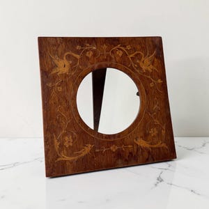 Op de afbeelding: Een vierkante houten fotolijst met een ronde opening in het midden. Het donkerbruine hout is voorzien van lichtgekleurde houten inlegwerkontwerpen van twee vogels die naar elkaar toe kijken, omringd door bloem- en rankmotieven aan alle zijden.