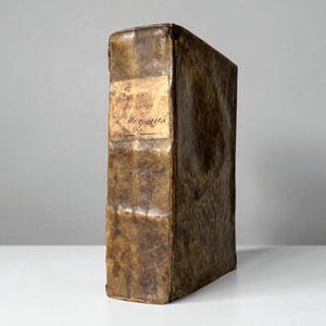 May include: A vintage, leather-bound book with a worn, brown cover. A rectangular label with faded text is affixed to the spine. The book is standing upright against a neutral background, showcasing its aged appearance and historical significance.