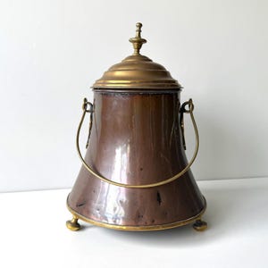 May include: A copper bucket with a brass lid, handle, and three small feet. The bucket has a tapered, cylindrical body with a smooth, metallic finish. The lid features a central finial, and the handle is attached to two side loops.