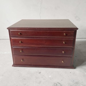 May include: A small, rectangular wooden chest with a dark reddish-brown finish. The chest features four horizontal drawers, each fitted with two small, circular brass-coloured knobs. The top surface is flat, and the unit has a structured, boxy design.