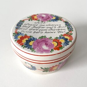 Op de afbeelding: Een ronde keramische pot met deksel, witte basis en rode rand. Het deksel is voorzien van een handgeschilderde bloemenkrans van roze rozen, blauwe korenbloemen en rode madeliefjes rondom de Duitse tekst: Hab' Sonne im Herzen, Und Butter auf's Brot, Und Geld zum Bezahlen, Dann hat's keine Not.