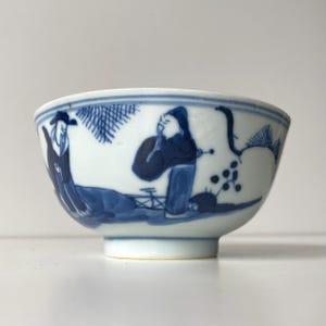 Peut inclure: Un bol en céramique blanc avec des illustrations bleues peintes à la main de personnages et de feuillages. Le bol a une forme arrondie avec un bord légèrement évasé et une petite base à pied. Les motifs bleus sont détaillés.