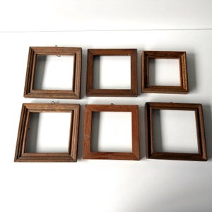 May include: A collection of six square wooden picture frames arranged in two rows. Each frame features a natural brown wood grain finish, a simple border design, and a small metal hanging loop attached to the top edge of the frame.