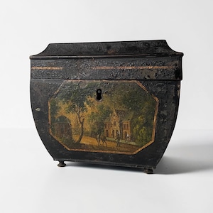 May include: Black antique box with a curved shape and small feet. The front features a painted landscape scene with a house, trees, and figures. The box has a keyhole and a gold-colored trim.