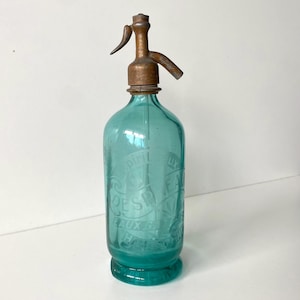 Op de afbeelding: Een vintage talingkleurige glazen seltzerfles met een verweerde metalen sifondop. Het glazen lichaam bevat geëtste tekst met "DESPREAUX" en "EAUX GAZEUSES" in een cirkelvormig ontwerp, met een doorzichtige interne buis zichtbaar in de fles.