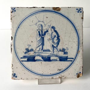 May include: A blue and white Delft tile depicting a scene from the Bible. Two figures, one with a halo, stand on a platform with a blue and white design. The tile is surrounded by a blue and white border.