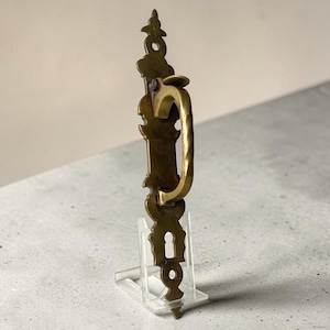 May include: A vintage brass door handle with an ornate design. The handle has a curved grip and decorative elements along its length, including a keyhole. The brass has a warm, aged patina, suggesting its antique nature. The handle is displayed on a clear acrylic stand.