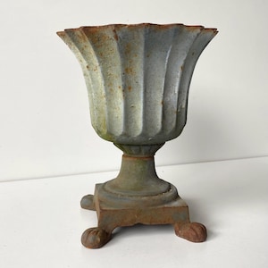 May include: A weathered cast iron urn with a fluted, scalloped rim and a grey finish showing patches of orange rust. The vessel features a central pedestal base supported by four distinct feet, displaying a textured, aged appearance throughout.