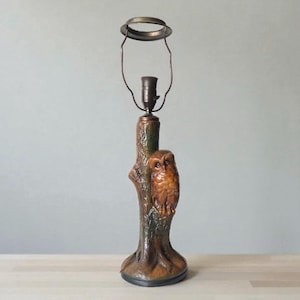 May include: A brown and green table lamp featuring a carved owl perched on a tree trunk. The lamp has a bronze-colored metal shade holder and a dark base. The owl is detailed with textured feathers.