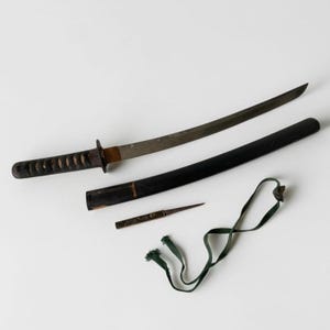 Puede incluir: Una espada katana japonesa con hoja de acero curva, mango envuelto en cordón negro y vaina oscura. Se incluye un pequeño cuchillo utilitario de metal y un cordón de tela verde. La hoja presenta grabados visibles a lo largo de la superficie metálica.