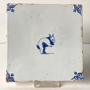 May include: Square white ceramic tile with a blue and white rabbit illustration. The rabbit is depicted in a jumping pose. Decorative blue corner accents complete the design. The tile has a rustic, aged appearance.